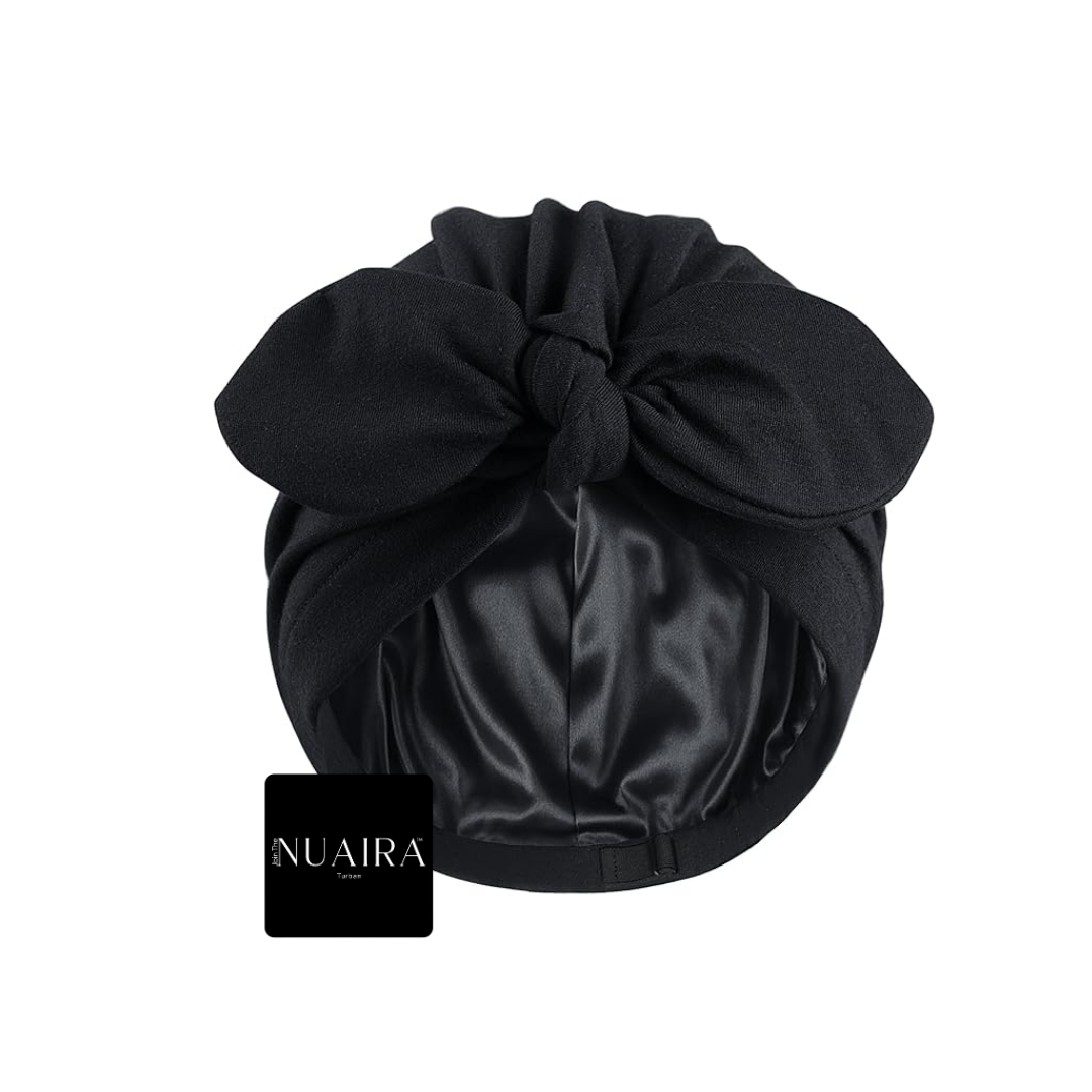A black hair wrap cap with a large knotted bow on top and a satin-like inner lining, shown against a plain white background. A small black brand tag reading NUAIRA is placed near the lower left of the product.