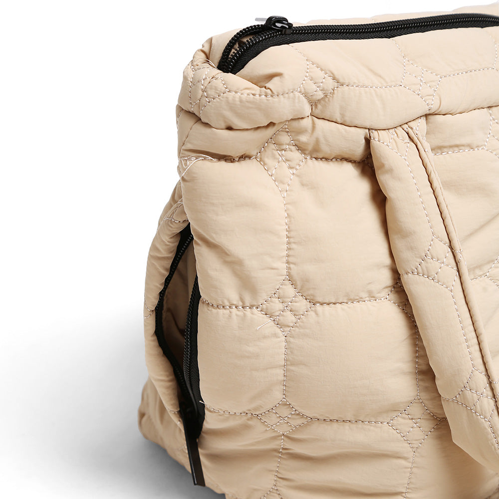 Close-up of a beige quilted puffer bag with black zippers, showing the texture and stitching details against a light gray background.