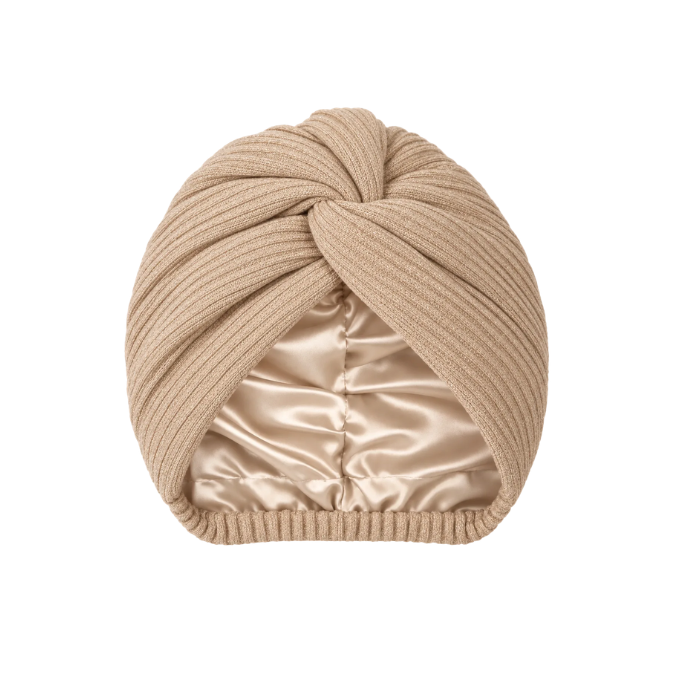 A beige ribbed knit turban hat featuring a central front twist detail. The interior is lined with a smooth, champagne-colored satin fabric. A small black rectangular tag with the white text "ALAIRA" is attached to the side edge.
