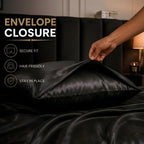 A close-up of a hand lifting a black satin pillowcase to demonstrate its envelope closure feature on a bed with matching black bedding. The left side of the image features text overlays explaining the benefits of the envelope closure: secure fit, hair friendly, and stay in place.