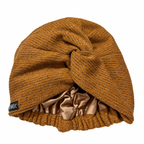 A mustard twisted knit turban-style beanie in a warm camel brown color, featuring a shiny satin inner lining and a small black brand tag on the side, set against a plain white background.