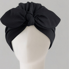 A black fabric headwrap turban hat with a large front bow, displayed on a white mannequin head against a light gray background.