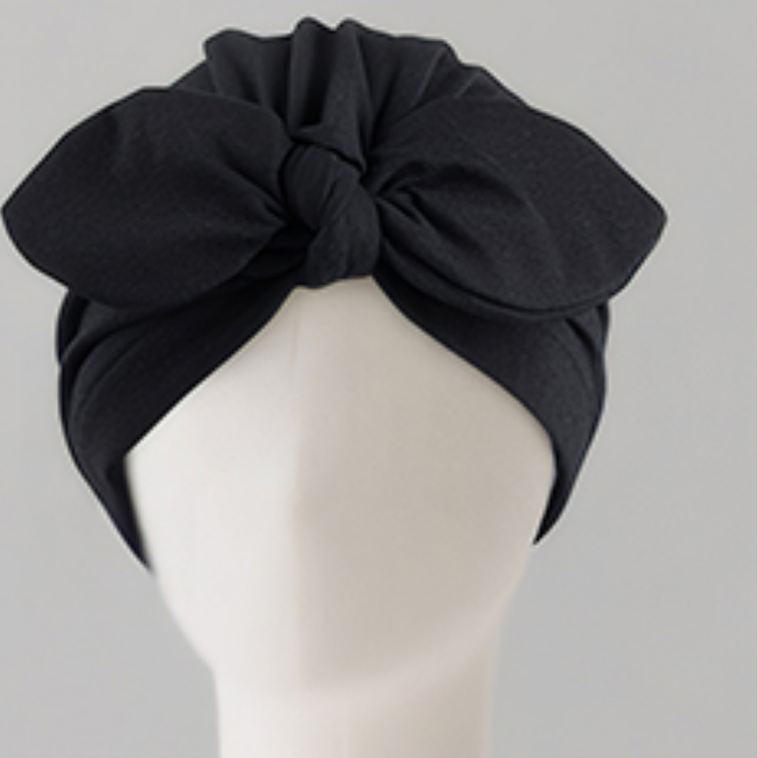 A black fabric headwrap turban hat with a large front bow, displayed on a white mannequin head against a light gray background.